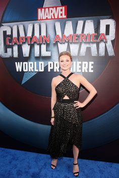 a woman in a black dress standing on a blue carpet with the captain america logo behind her