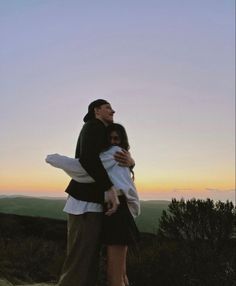 a man and woman embracing each other on top of a hill at sunset with the sky in the background