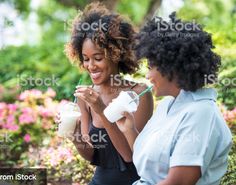 two women are drinking milkshakes outside in the day time and smiling at each other