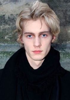 a young man with blonde hair and blue eyes wearing a black coat looking at the camera