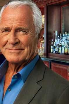 an older man wearing a suit and blue shirt in front of a bar with liquor bottles