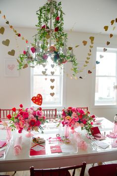 the table is set for valentine's day with pink flowers and hearts hanging from the ceiling