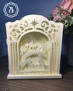 a paper cut nativity scene is displayed on a wooden table next to a vase with flowers