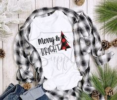 a white shirt with merry and bright on it next to pine cones, plaid shirts and jeans