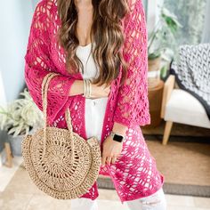 a woman wearing a pink crochet cardigan holding a straw bag and smiling