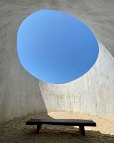 a bench sitting under a blue sky next to a cement wall with a circular hole in it