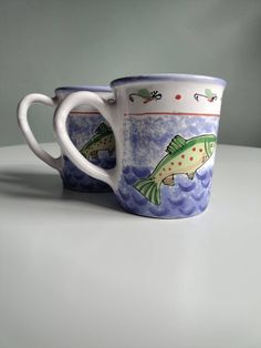 two coffee mugs with fish painted on them sitting side by side in front of a gray background