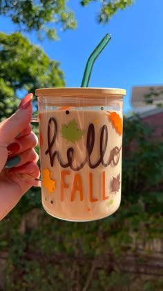 a hand holding up a glass with hello fall written on it and leaves in the bottom