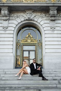 a man and woman sitting on steps in front of a city hall building with the doors open