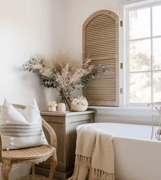 a white bath tub sitting under a window next to a wooden chair with pillows on it