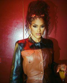 a woman in a leather jacket holding a glass and looking at the camera while standing next to a red wall