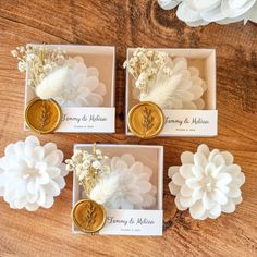 three wedding favors in boxes with white flowers and gold buttons on them, sitting on a wooden table