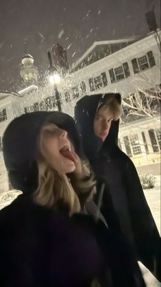 two people standing in front of a building with snow falling on them and one person wearing a hat