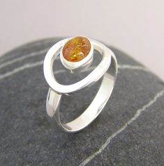 This modern statement ring features a glowing Baltic amber cabachon caught in an orbit of sterling silver. The band measures just under 1/4". The stone is 8 x 10 mm.All of our pieces are hand wrought not cast, making each unique.Comes packaged in a soft pouch in a gift box tied with ribbon, ready for giving!Amber is the fossilized resin of ancient evergreen trees.  It is said to help manifest desires, promote clarity of thought and wisdom. It is reputed to draw out negativity, and bring balance, Modern Amber Oval Ring, Modern Amber Rings For Anniversary, Amber Sterling Silver Oval Cabochon Ring, Amber Oval Cabochon Sterling Silver Ring, Amber Sterling Silver Ring With Oval Cabochon, Orbit Ring, Pmc Jewelry, Yellow Jewelry, Silver Jewelry Design