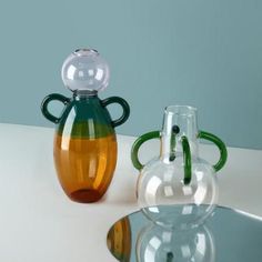 two glass vases sitting next to each other on a white counter top, one with green handles