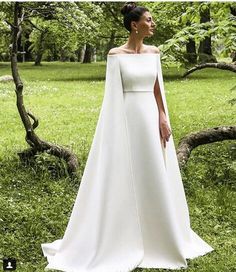 a woman in a white wedding dress standing in the grass with her cape over her shoulders