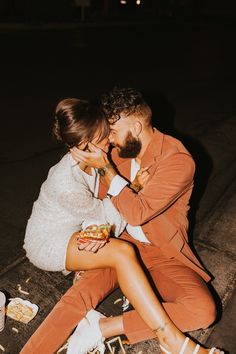 a man and woman sitting on the ground sharing a kiss while eating food at night