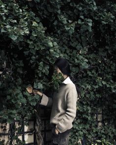 a woman standing in front of a fence next to a green bush with leaves on it