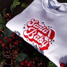 a white t - shirt with the words santa claus on it next to pine cones