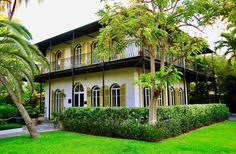 a large white house with yellow shutters and trees in the front yard, surrounded by lush green grass