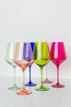 five different colored wine glasses lined up in a row on a white surface, one is empty