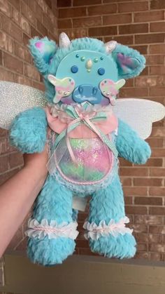 a hand holding a blue teddy bear in front of a brick wall with a pink bow on it's chest