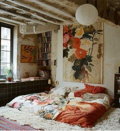 an unmade bed in front of a window with bookshelves and paintings on the wall