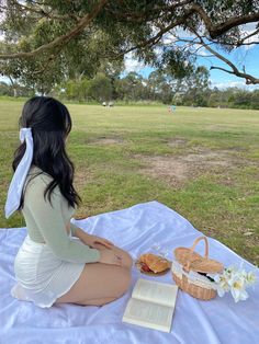 a woman sitting on a blanket with a picnic basket and book in front of her