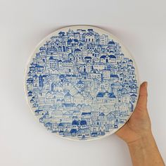 a hand holding up a blue and white plate with buildings on the outside, in front of a white background