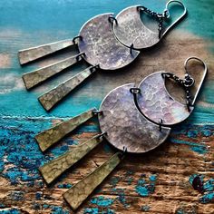 two silver and bronze earrings on top of a piece of blue wood with chains hanging from it