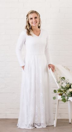 a woman standing in front of a white chair wearing a white dress and smiling at the camera