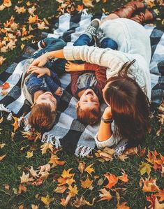 three people laying on top of a blanket in the grass with leaves all around them