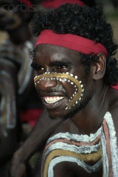 an african man with painted face and headdress smiles at the camera while sitting in front of other men