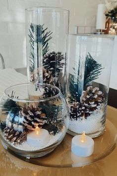 two clear vases filled with pine cones and candles on a glass table next to a candle holder