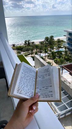 someone is holding an open book in their hand near the ocean and buildings with palm trees