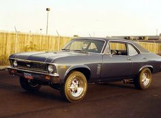 an old muscle car parked in a parking lot next to a wooden fence and building