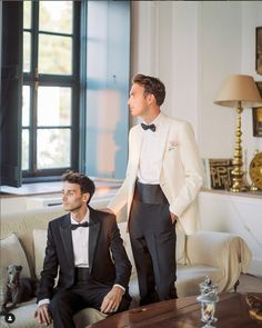 two men in tuxedos are sitting on a couch and one man is standing