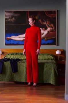 a woman standing in front of a bed wearing a red jumpsuit and matching heels