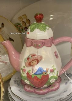 a tea pot with a strawberry on it sitting next to other plates and dishes in the background