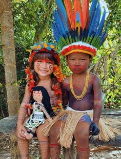 two young children dressed in native american clothing and headdress sitting on a tree stump