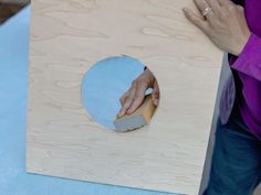 a person holding a piece of wood in front of a circular hole on the side of a table