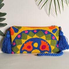 a colorful clutch bag with tassels on the front and side, sitting next to a potted plant