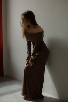 a woman leaning against a wall in a long brown dress with her hands on her hips