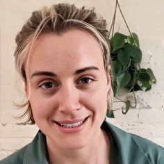 a woman with blonde hair and green shirt smiling at the camera while standing in front of a white brick wall