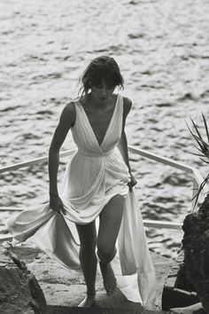 black and white photograph of woman in dress walking on pier next to body of water