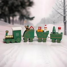 a green toy train traveling down a snow covered road next to a christmas tree and small house