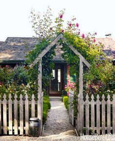 a white picket fence with flowers growing over it and a house in the background,