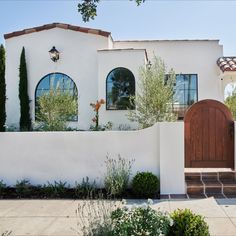 a white stucco house with wooden door and arched windows, surrounded by greenery on either side