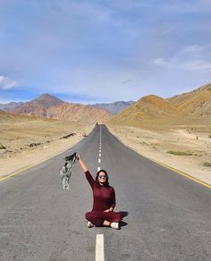 a woman sitting on the side of an empty road with her arms in the air