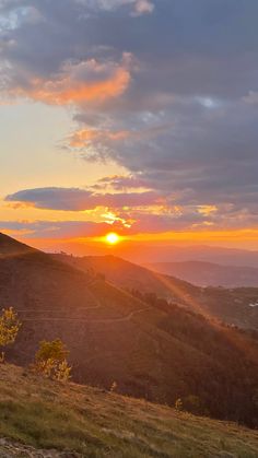 the sun is setting on top of a hill with hills in the foreground and clouds in the background